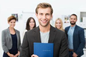 Jeune homme souriant, candidat à un emploi, tenant un dossier bleu avec son curriculum vitae, posant devant ses nouveaux collègues de travail ou son équipe.jpeg